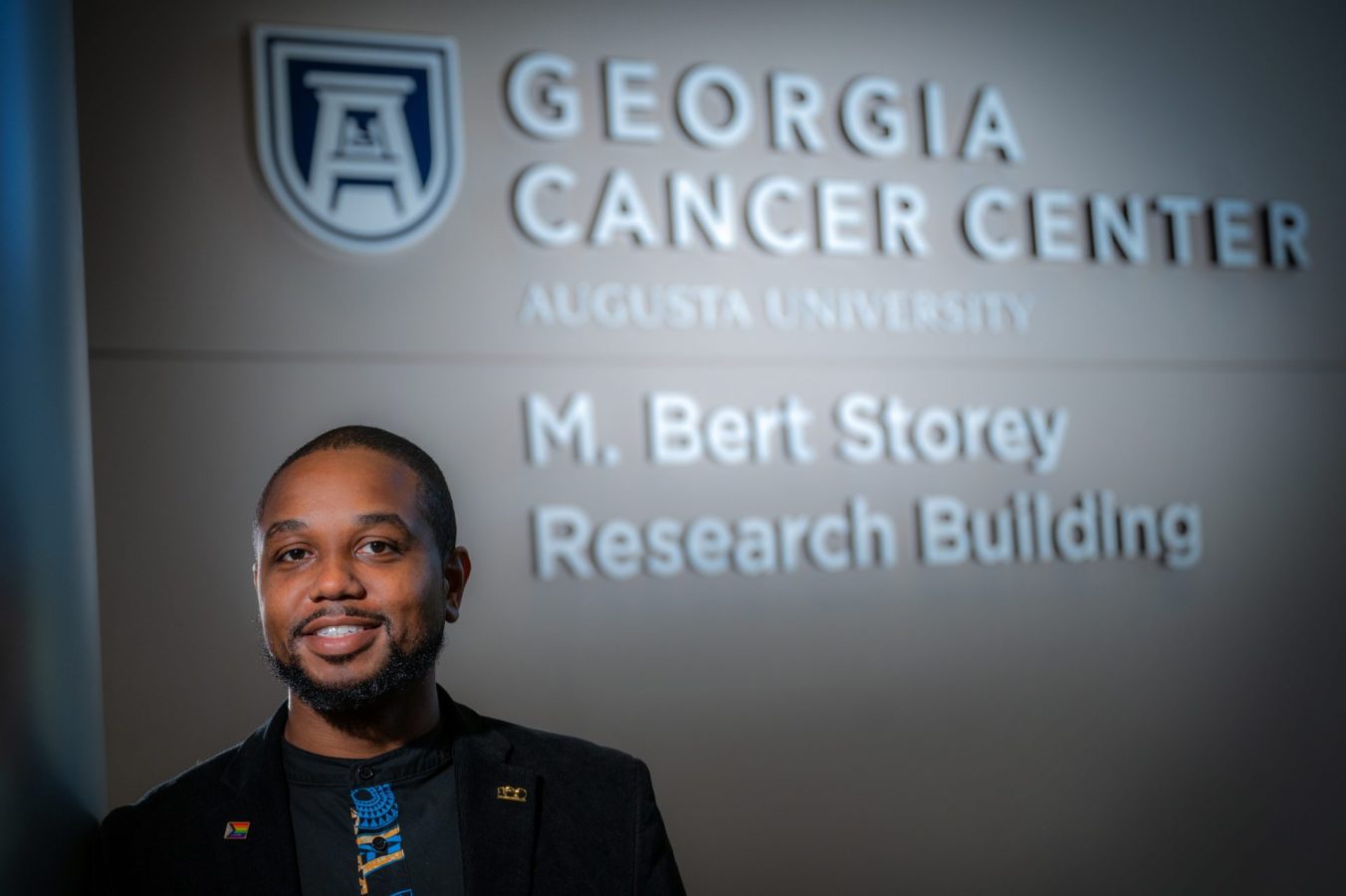 Man in black coat and blue and black shirt stands in front of sign that reads "Georgia Cancer Center"