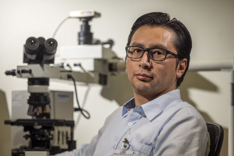 man with glasses in blue shirt sits in front of microscope