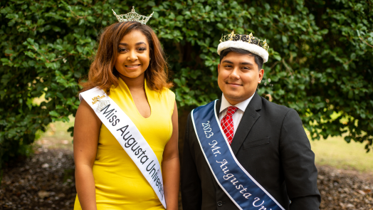 a man and a woman stand outside, wearing crowns and sashes