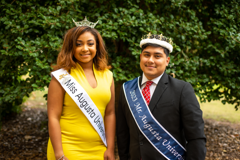 a man and a woman stand outside, wearing crowns and sashes