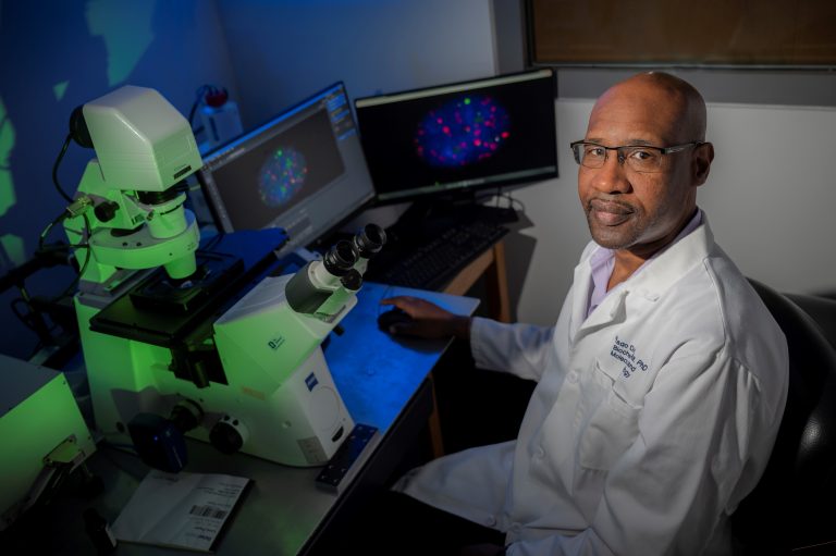 Man in glasses sits in front of microscope and looks at camera