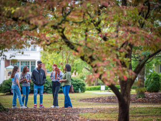 group of five college students stand together outside under trees
