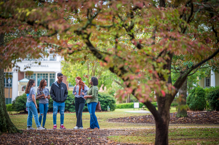 group of five college students stand together outside under trees