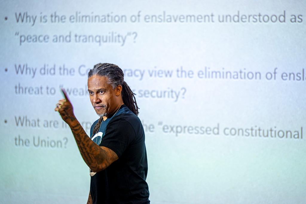 man speaking to audience in front of a projector screen