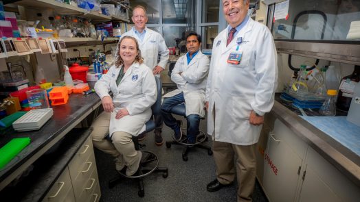 Four people in white lab coats stand in lab and smile at camera