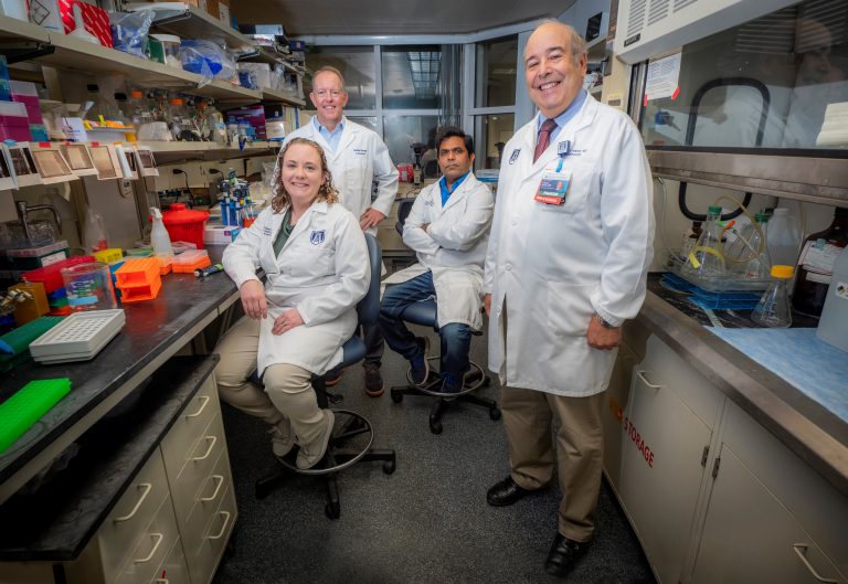 Four people in white lab coats stand in lab and smile at camera