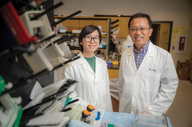 Man and woman in white coats stand in front of microscope in large lab