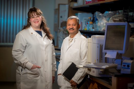 Man and woman in white coat stand in lab in front of computer screen