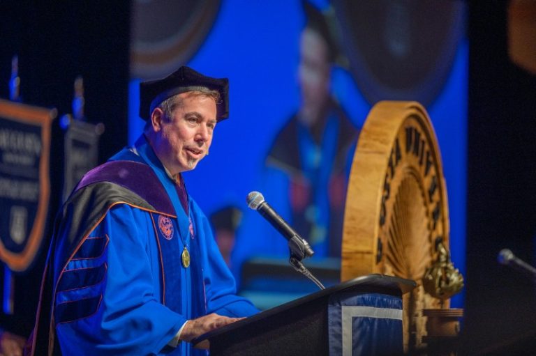man wearing higher education graduation regalia speaks at a podium