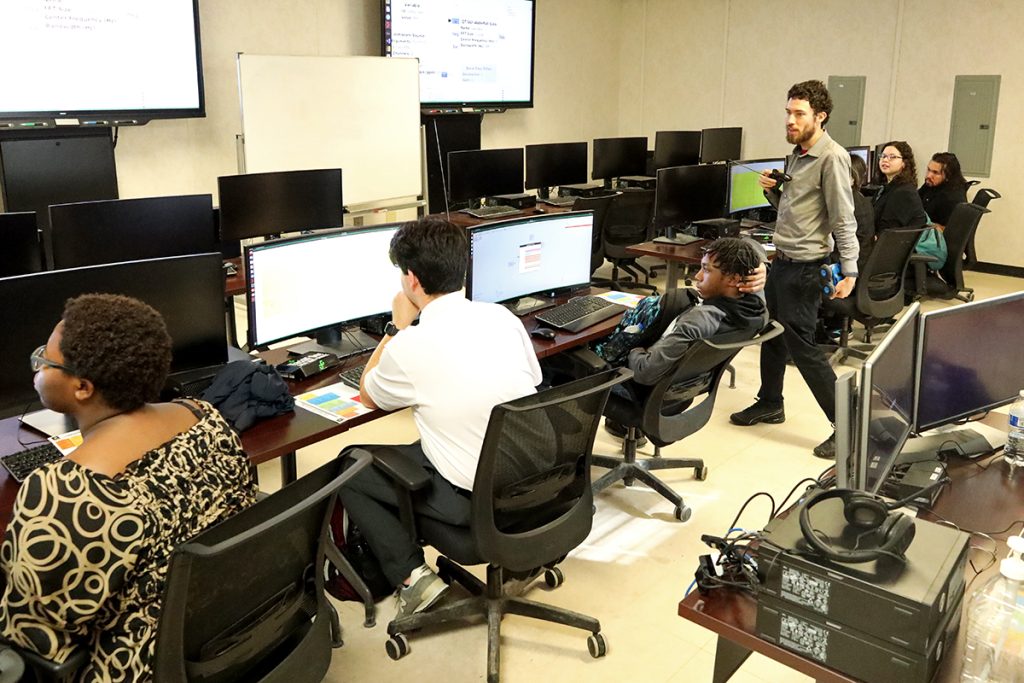 Six people sit at computers in a computer lab while an instructor holding a remote control and remote control car walks around the classroom