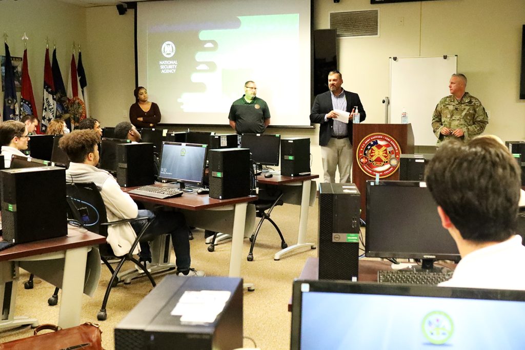 Three men and one woman stand in front of a group of people in a computer lab during a presentation.