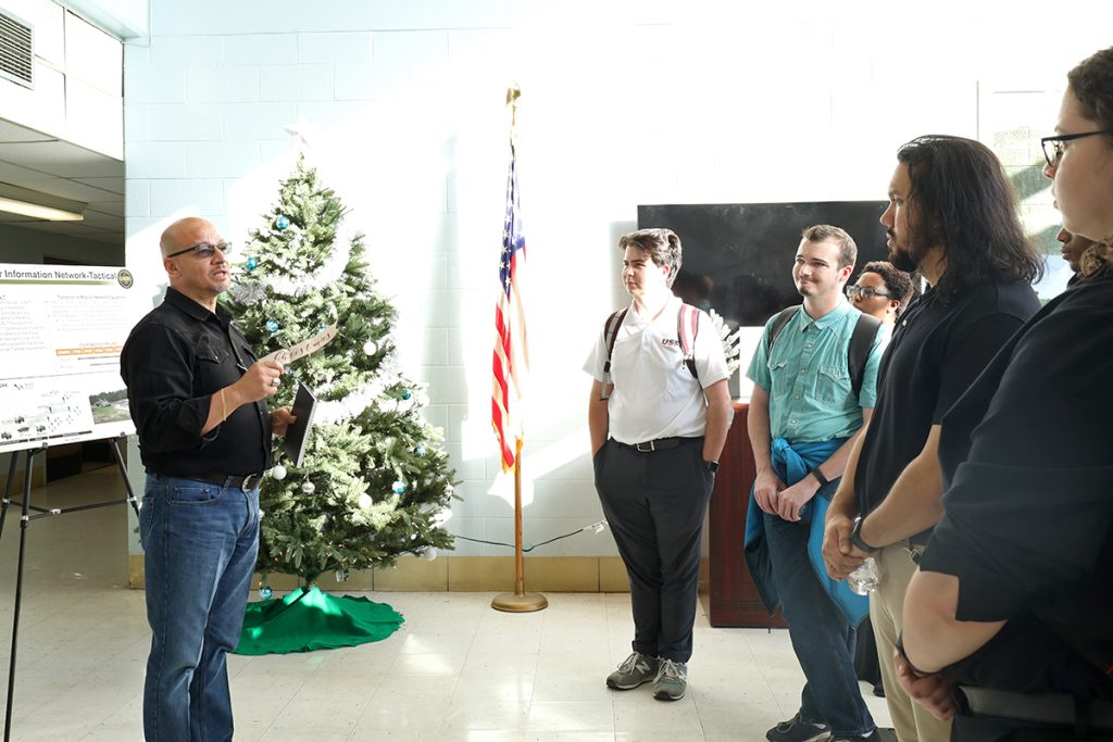 A man talks to a group of students standing in the lobby of a building with a Christmas tree and American flag in the background.