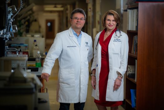 Man and woman, both in white coats, stand in hospital hallway and smile at camera