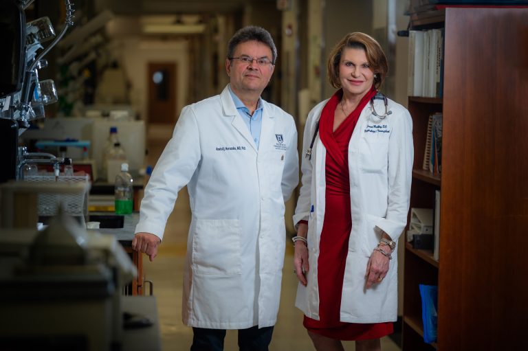 Man and woman, both in white coats, stand in hospital hallway and smile at camera