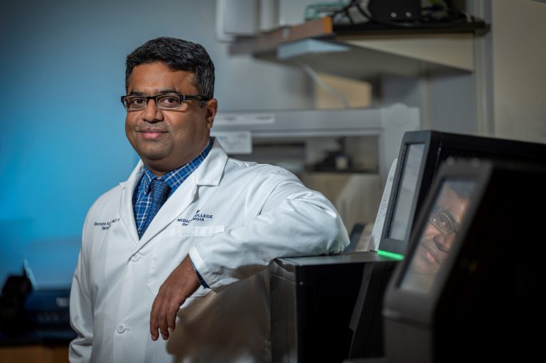 Man in white coat and blue shirt smiles at camera with arm propped on lab equipment