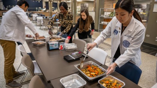 Students stand around table spooning food into containers