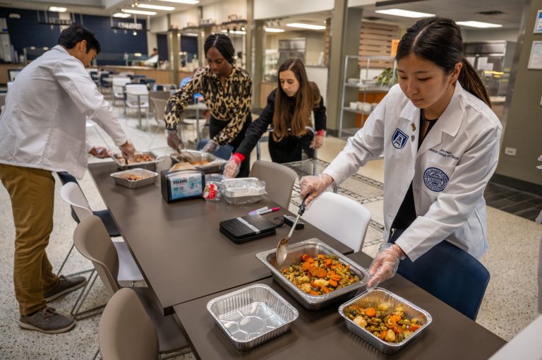 Students stand around table spooning food into containers
