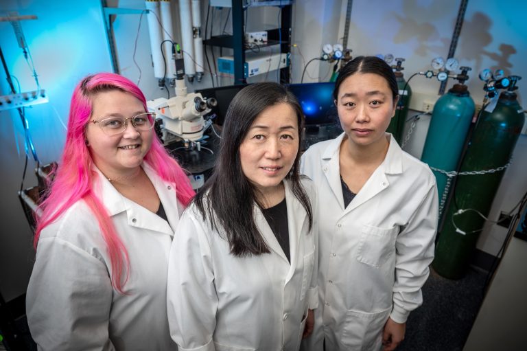 Three women stand in white coats with lab in background.