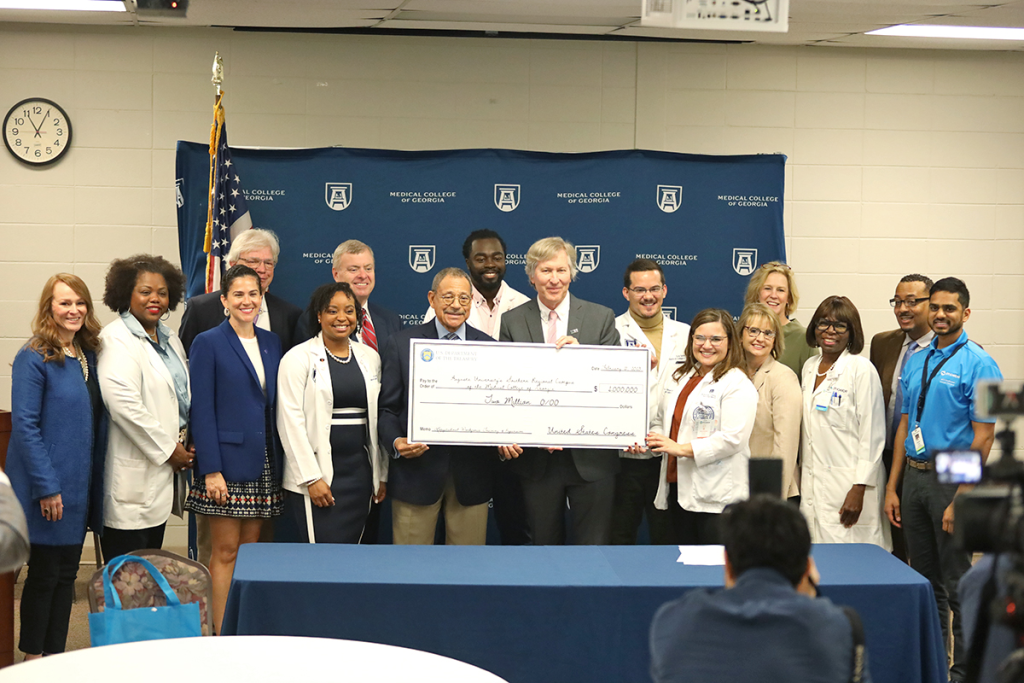 A large group of men and women gather around a large commemorative check while people take their photo