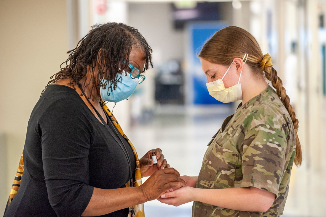 An African American woman holds a white woman's hands as they bow their heads in prayer. Both are wearing masks in a hospital setting.