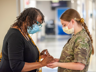 An African American woman holds a white woman's hands as they bow their heads in prayer. Both are wearing masks in a hospital setting.