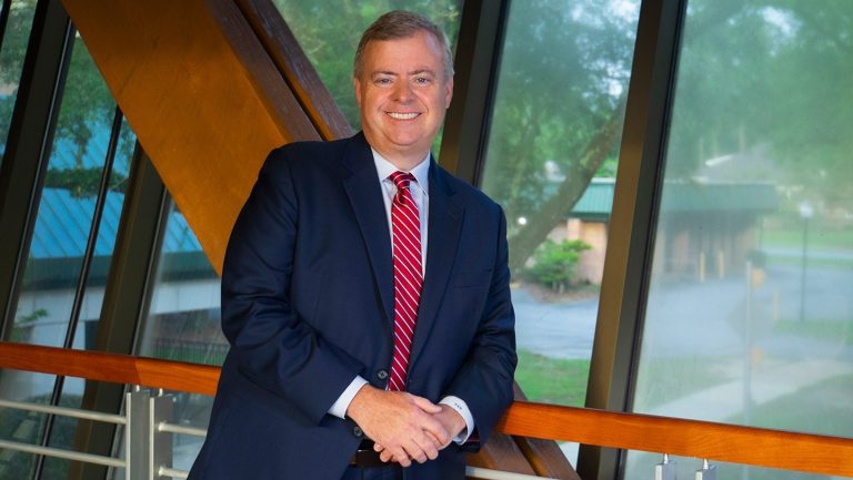 Man in blue suit and red tie leans against wooden beam