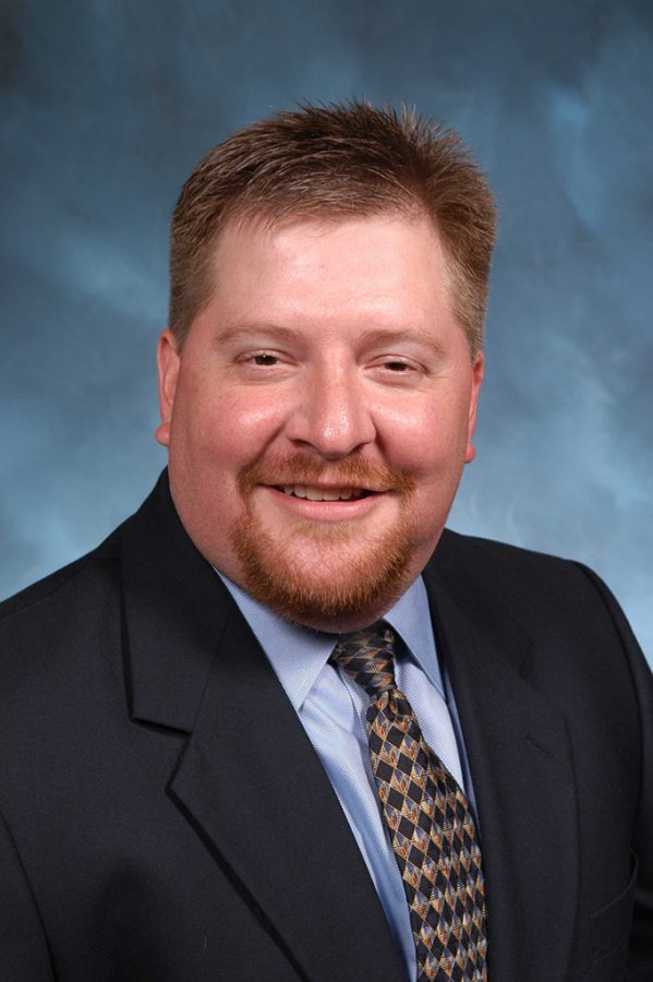 Man in a black suit jacket, light blue shirt and patterned tie smiles at the camera