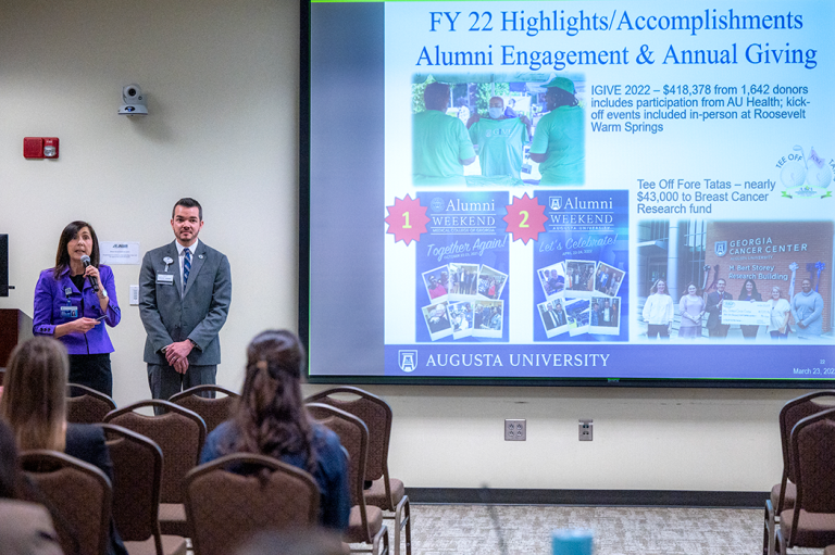 A man and woman stand in front of a projection screen, presenting to a room full of people