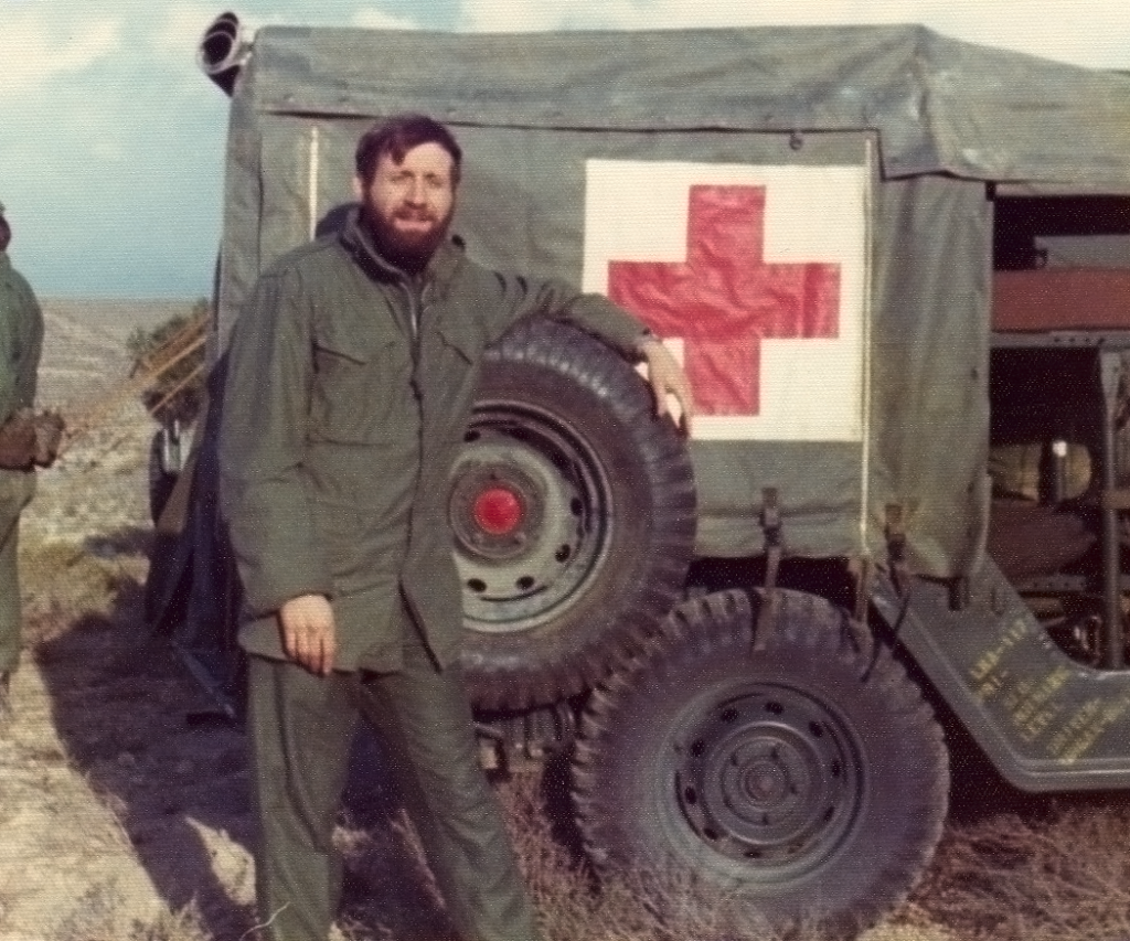 man with a beard stands in front of a military vehicle with the markings of a hospital's red cross and white background