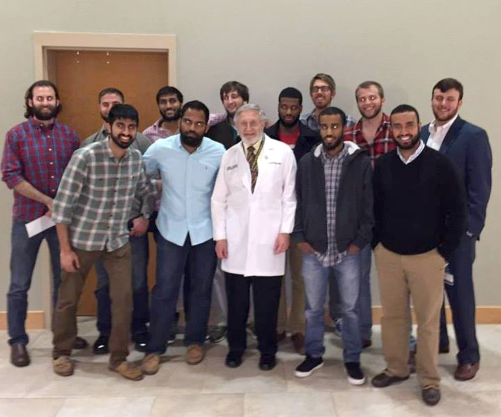 older man in lab coat stands with 12 students for a photo