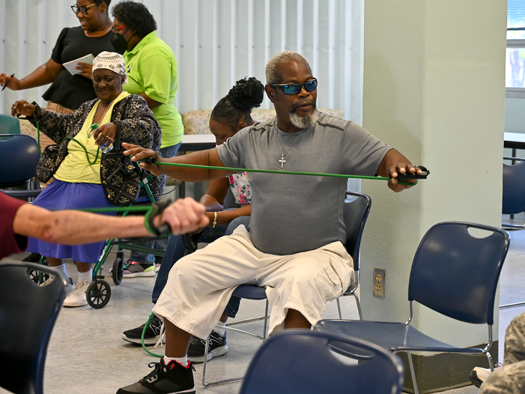 man wearing sunglasses sits in a chair and uses a tension rubber band to exercise his arms