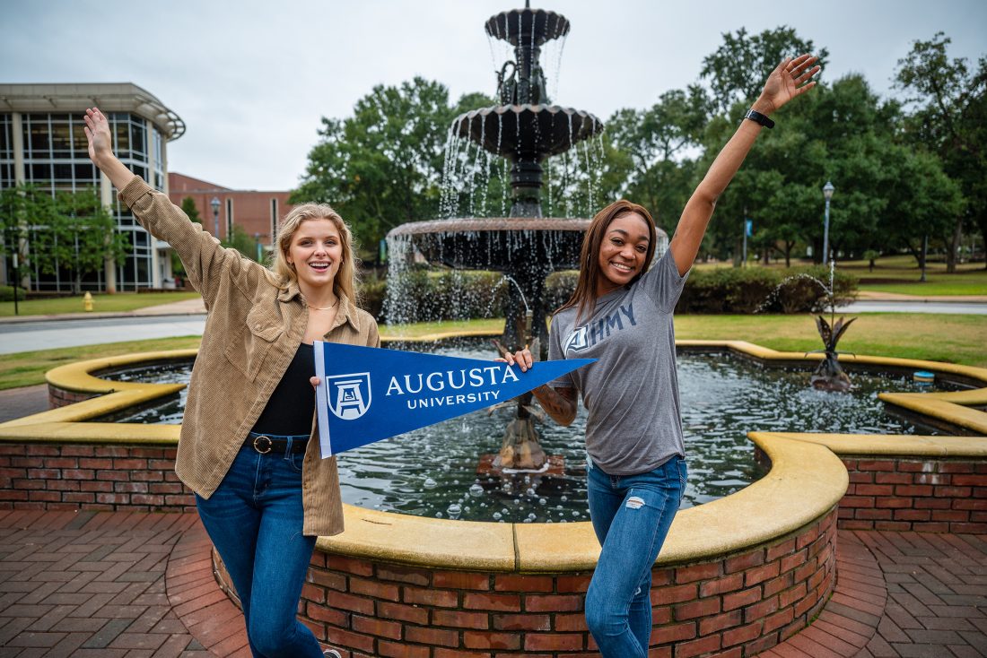 Two women hold Augusta University pennant in front of fountain