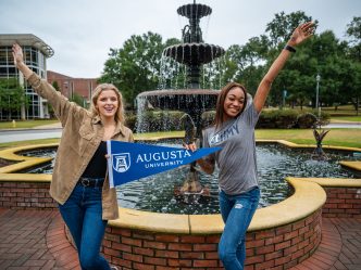 Two women hold Augusta University pennant in front of fountain