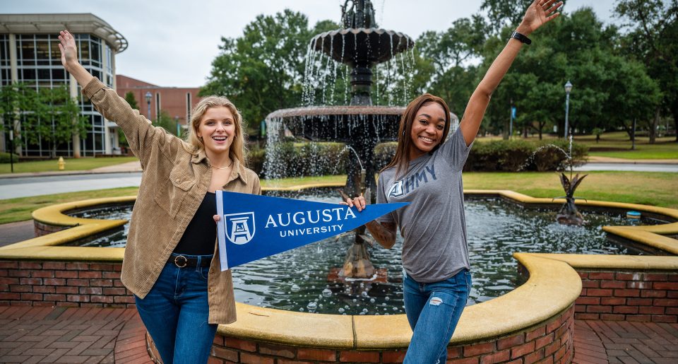 Two women hold Augusta University pennant in front of fountain