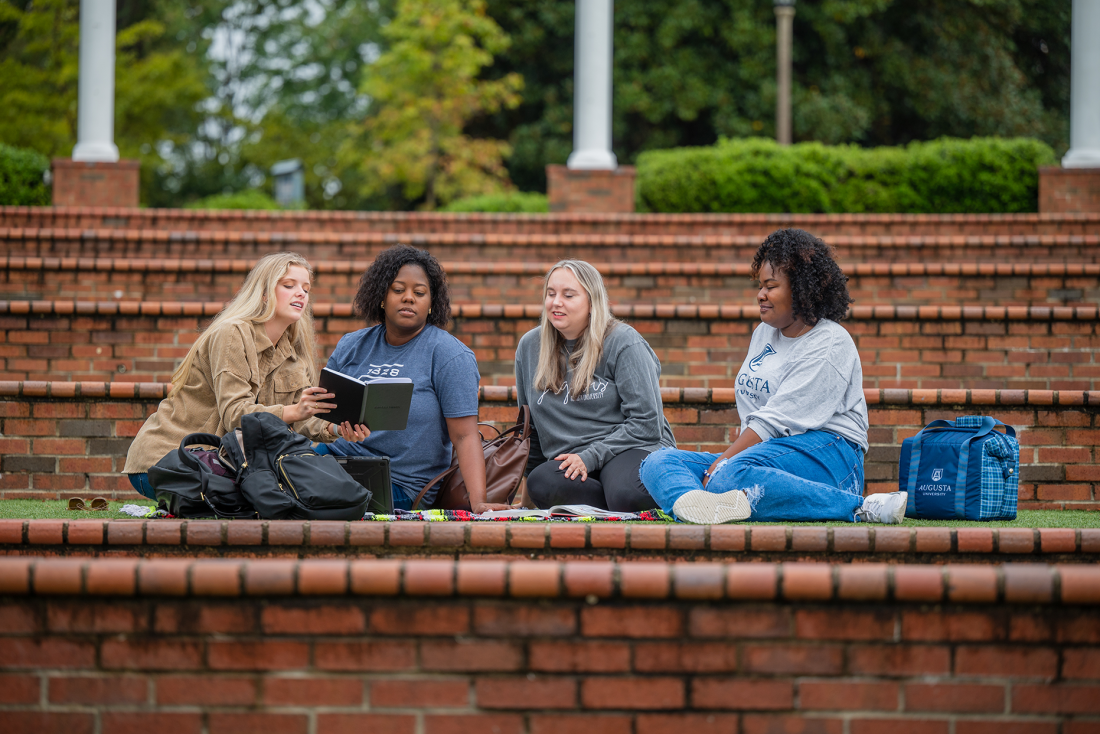 four women sitting in amphitheater talking and looking at a book