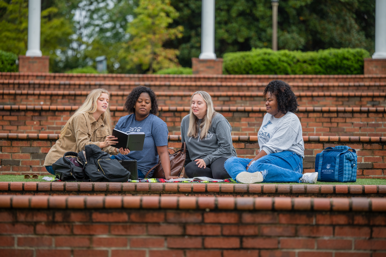 four women sitting in amphitheater talking and looking at a book