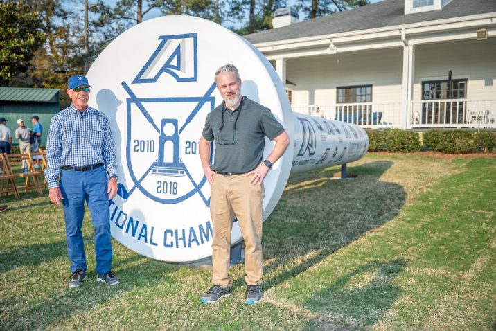 two men stand in front of an oversized golf tee sculpture