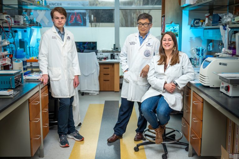Three people in white coats stand in lab looking at camera