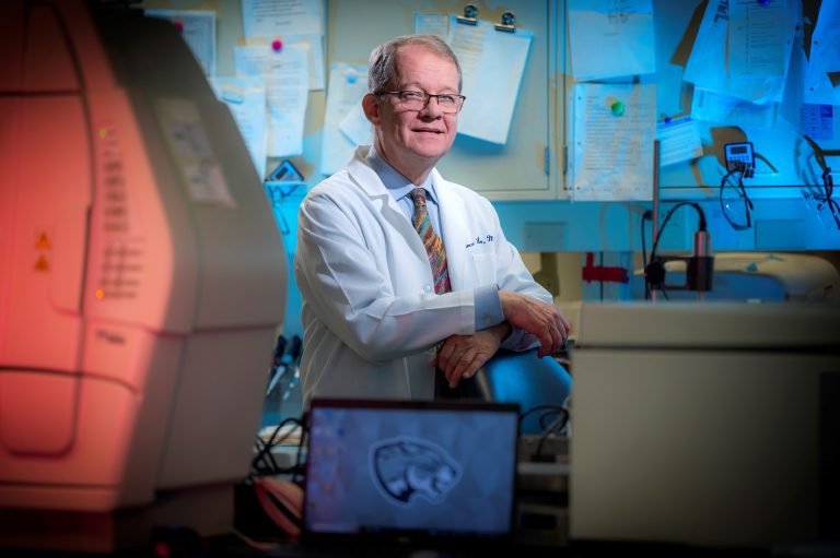 Man in white lab coat and glasses props hands on lab equipment
