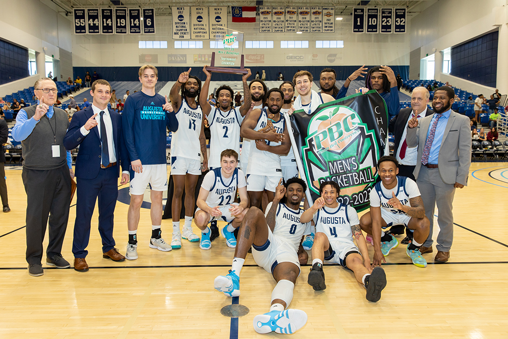 Men's Basketball team celebrating their victory while holding a trophy and banner