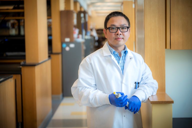 Man with glasses, blue shirt, white coat, wearing glvoes, stands with lab in background