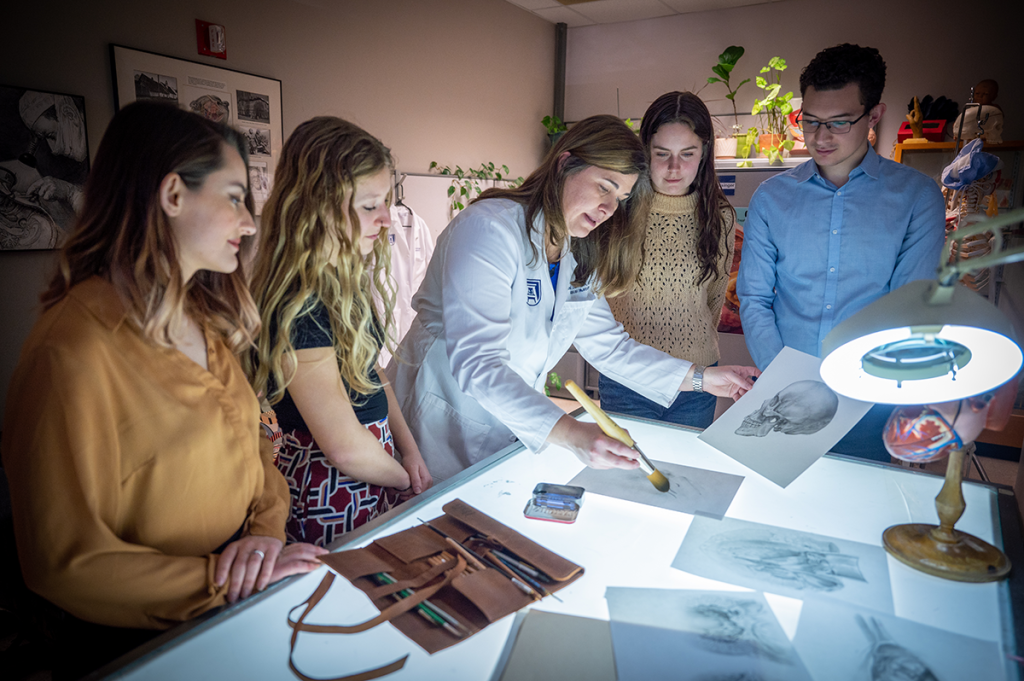 four women and one man gather around a table looking at drawings of human anatomy