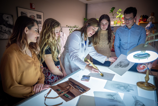 four women and one man gather around a table looking at drawings of human anatomy