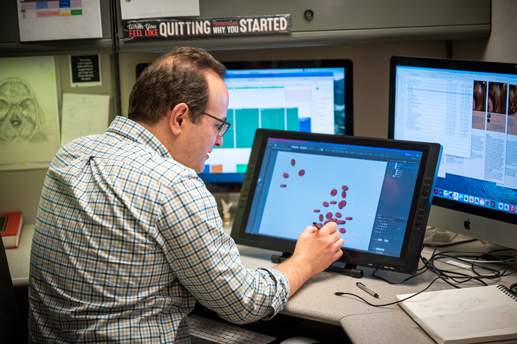 man sits at a desk with three computer screens, using a tablet and stylus to draw on one of the screens.