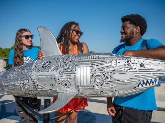 Three students standing behind a metal shark sculpture.