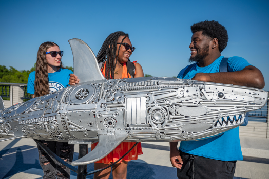 Three students standing behind a metal shark sculpture.