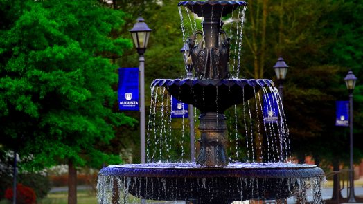 A fountain flows with a blue Augusta University banner hanging off a light pole in the background