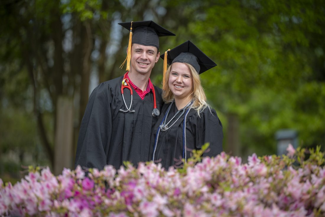Man and woman in cap and gown behind azalea bush