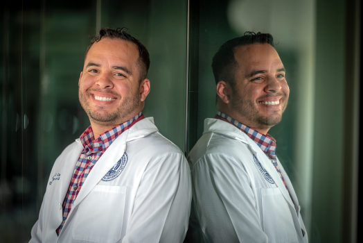 man wearing a health care professional coat leans against a window and smiles