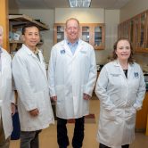 Five people in white coats stand in middle of lab